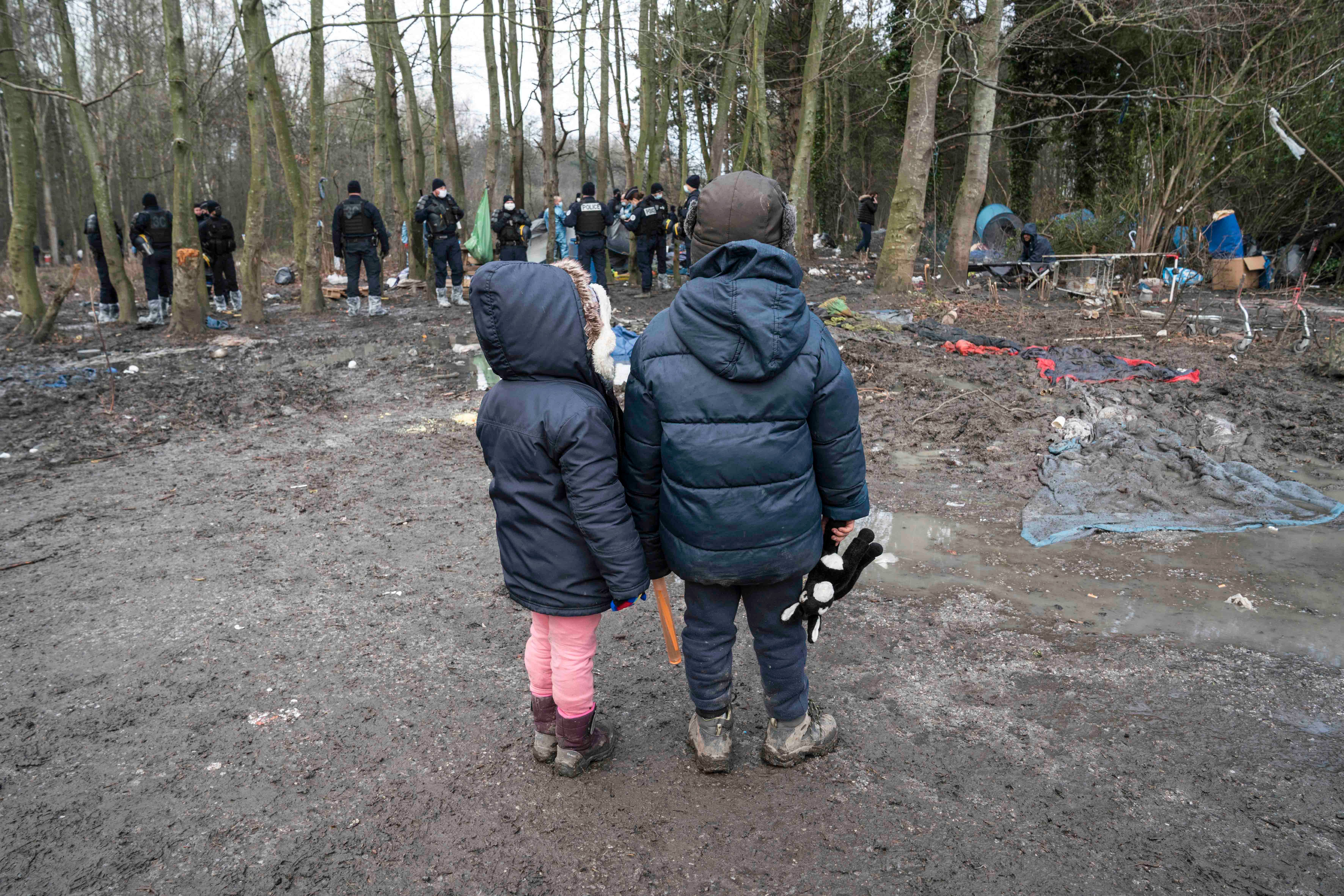 Two children wearing winter coats stand in a forest in front of a group of police officers