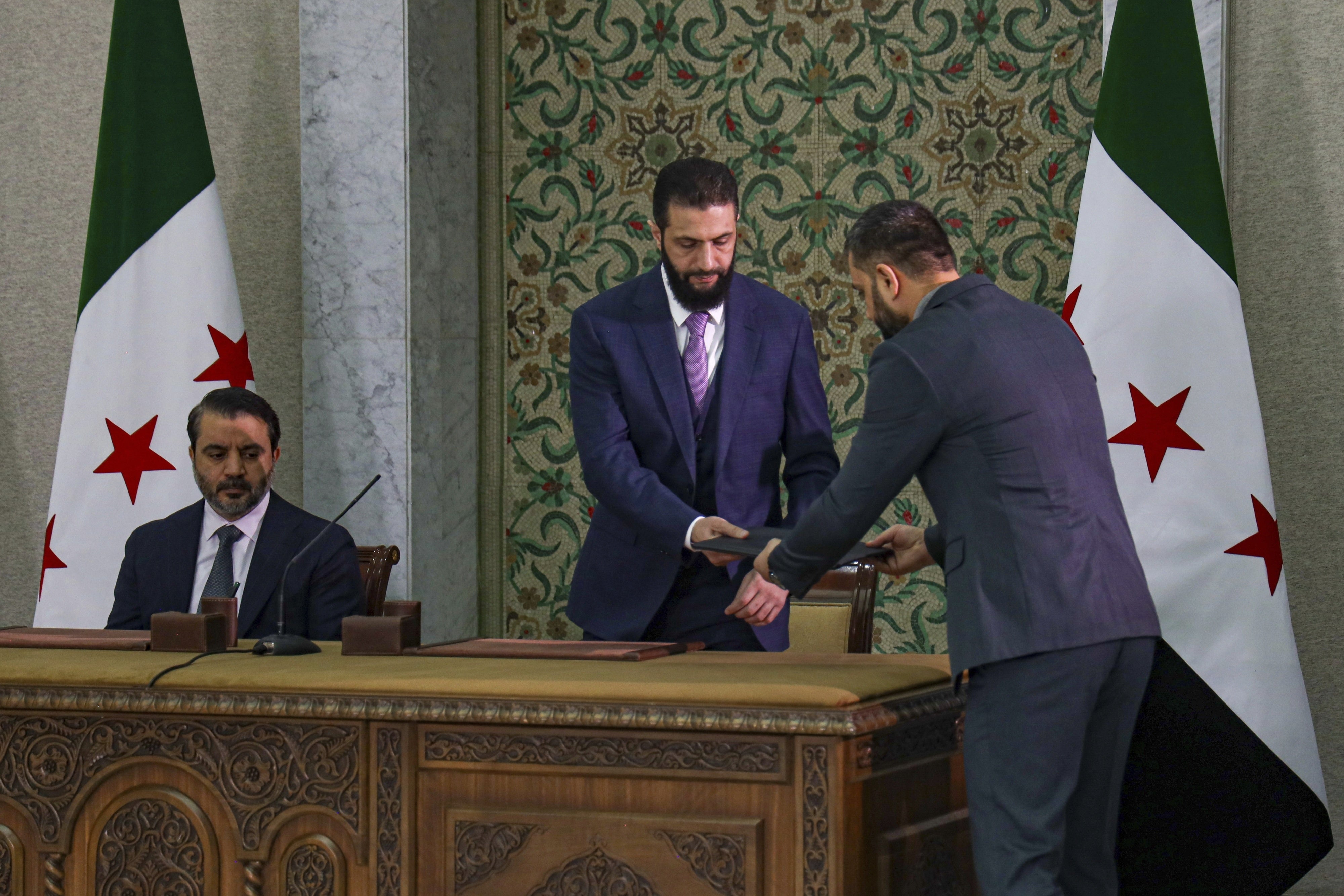 الرئيس السوري المؤقت أحمد الشرع (وسط) يتحضر لتوقيع الإعلان الدستوري السوري في دمشق، 13 مارس/آذار 2025. 