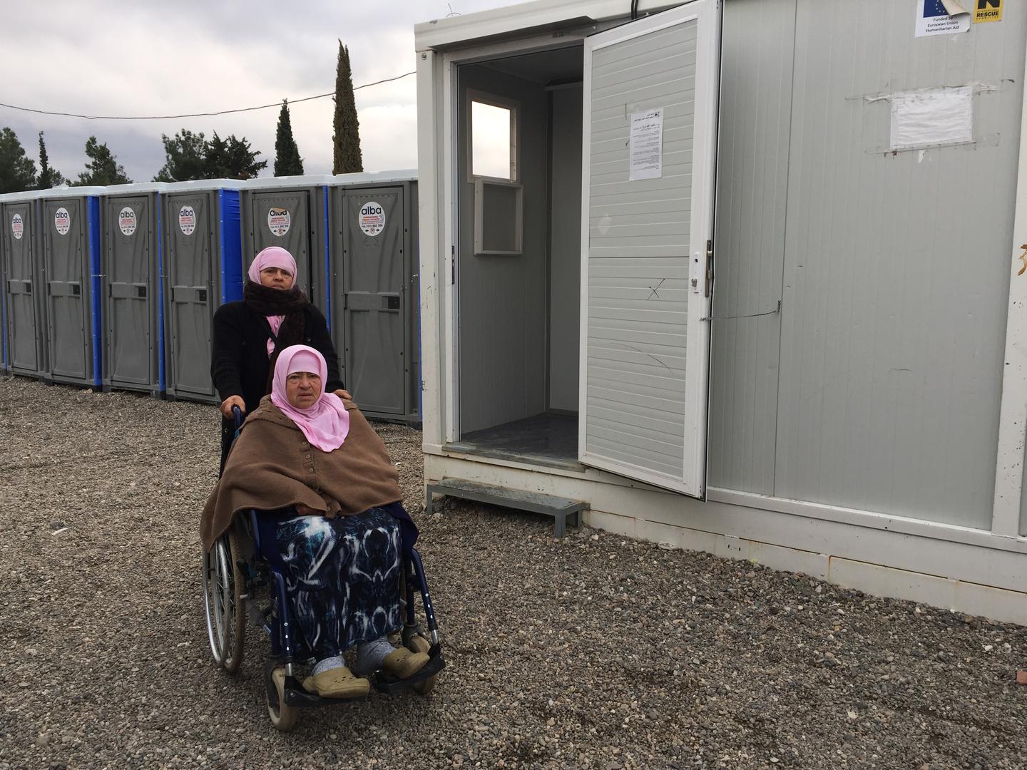 Naima, 70, an older woman with a disability from Aleppo, Syria, with her daughter Hasne, in front of the shower area in Cherso camp, Thessaloniki. The showers are not accessible for people who use a wheelchair.