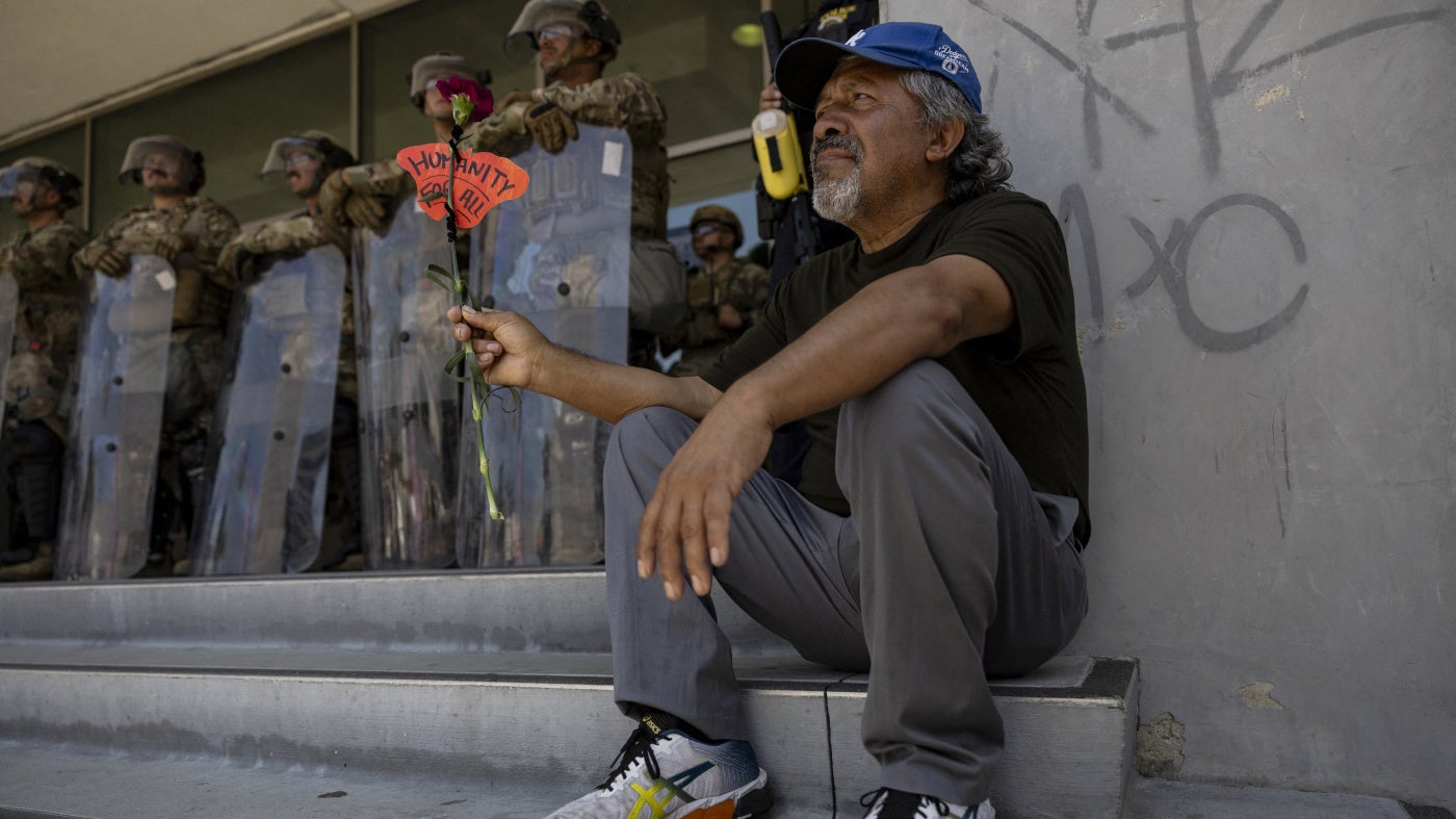 A man holds a flower and the message "Humanity for All" in front of a line of soldiers