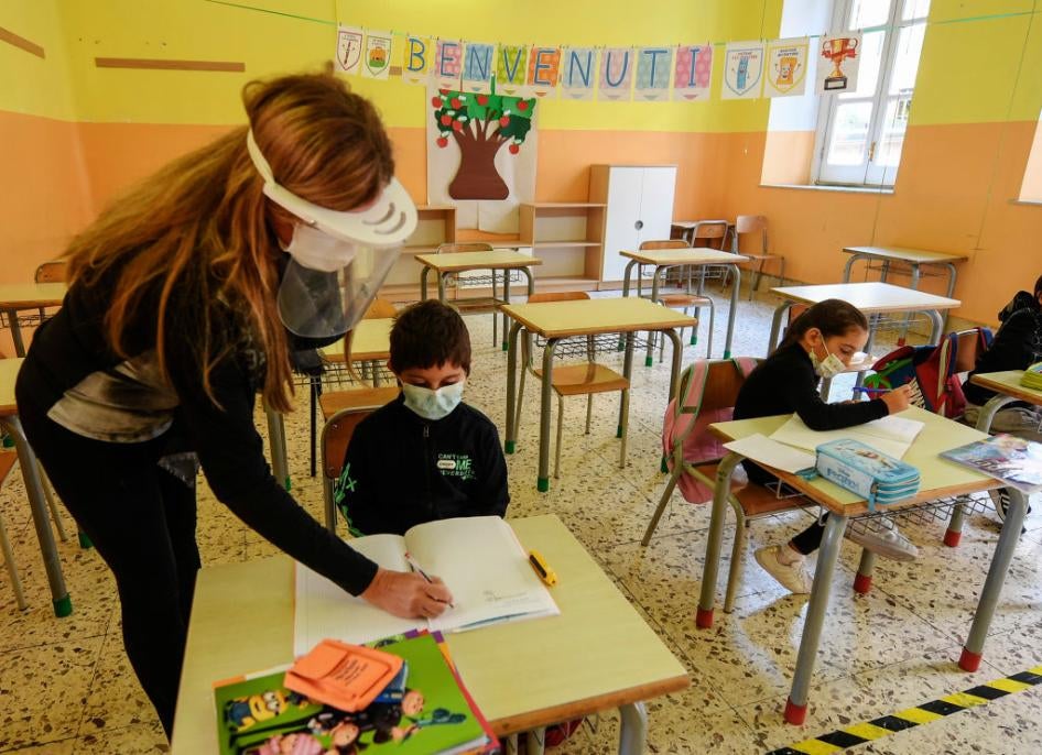 Teacher supports three students sitting in an otherwise empty classroom, all wearing face masks. 