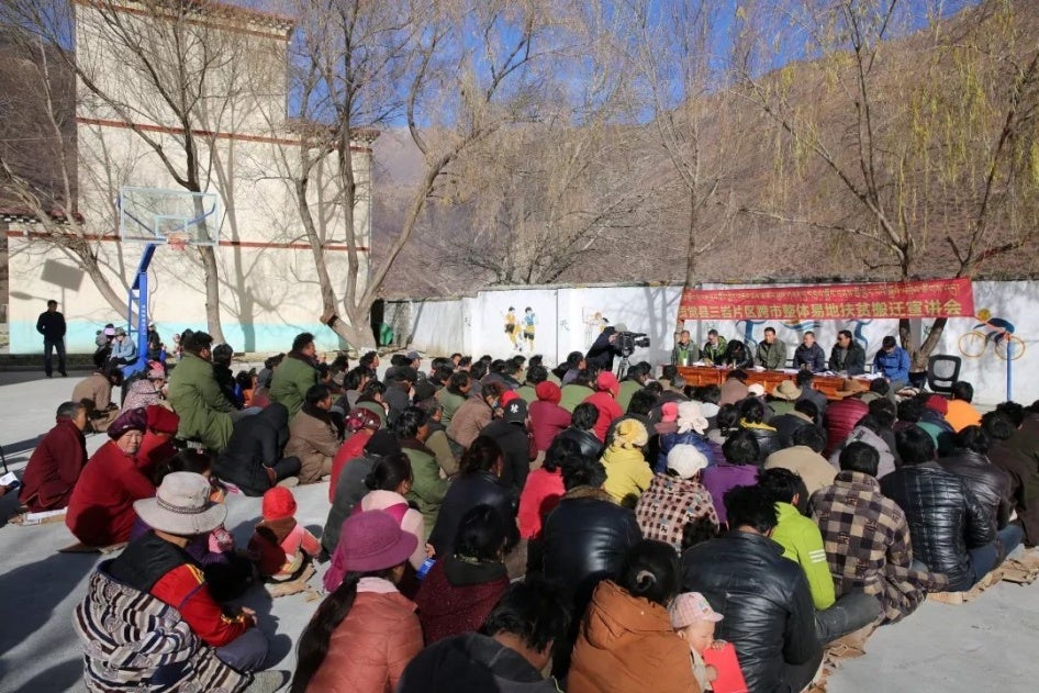An outdoor meeting in a town with a group of people seated on the ground in front of a table where officials are presiding