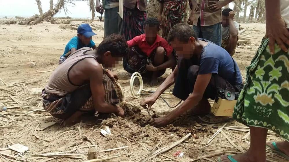 A deminer lacking protective equipment works to defuse a mine, with children and what appear to be other civilians watching.