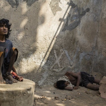 A man sitting on a concrete structure with a chain around his ankle. Another man is lying on the concrete floor.